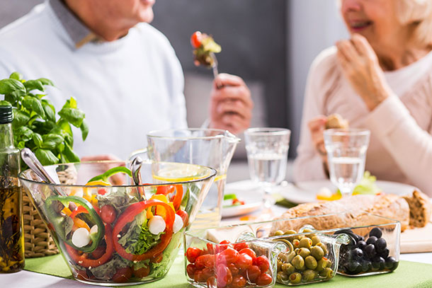 Ein frischer knackiger Salat und Antipasti stehen auf einem Tisch. Ein älteres Paar geniesst das Essen im Hintergrund. 