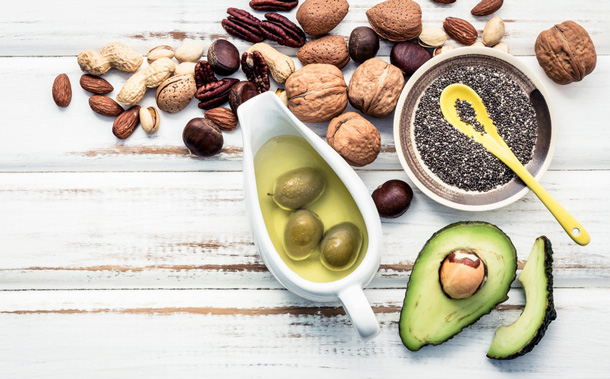 Im Bild sieht man Chia-Samen und Olivenöl in je einer kleine Schüssel. Daneben liegen eine aufgeschnittene Avocado und unterschiedliche Nüsse.