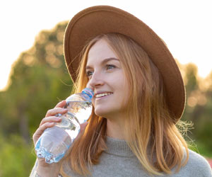 Eine Frau steht im Freien und möchte gerade frisches Wasser trinken. Sie hält eine PET-Flasche mit Wasser in der Hand.