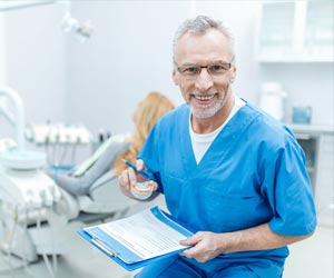 Ein Zahnarzt in seiner Praxis. Er sieht freundlich in die Kamere und hält Papier, Stift und eine Zahnprotese in der Hand.