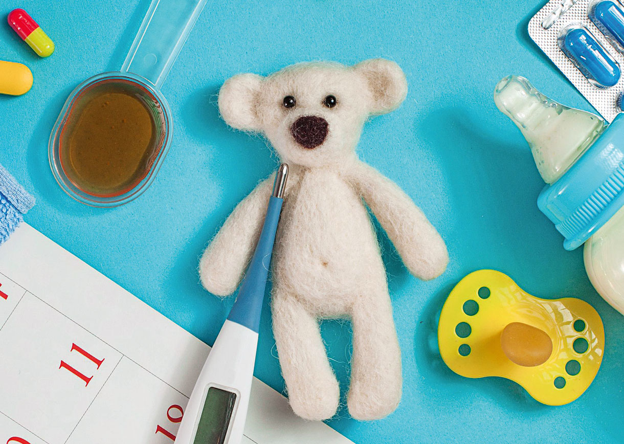 Diverse Dinge liegen auf blauem Hintergrund. Ein kleiner weisser Teddy, Schnuller, Tabletten, Fiebermesser und ein Plastiklöffel mit Hustensaft. Sinnbild für das Sammelsurium, wenn man ein krankes Kind hat.